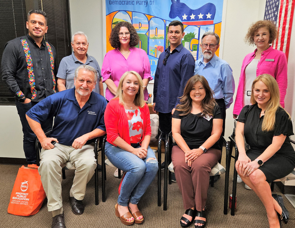 Group portrait of ten adults attending a Democratic Party of Orange County candidate training event. Five people stand and five sit in front of a DPOC banner and American flag inside a meeting room. Participants wear business-casual attire and smile toward the camera, with campaign and education-related materials visible in the room.