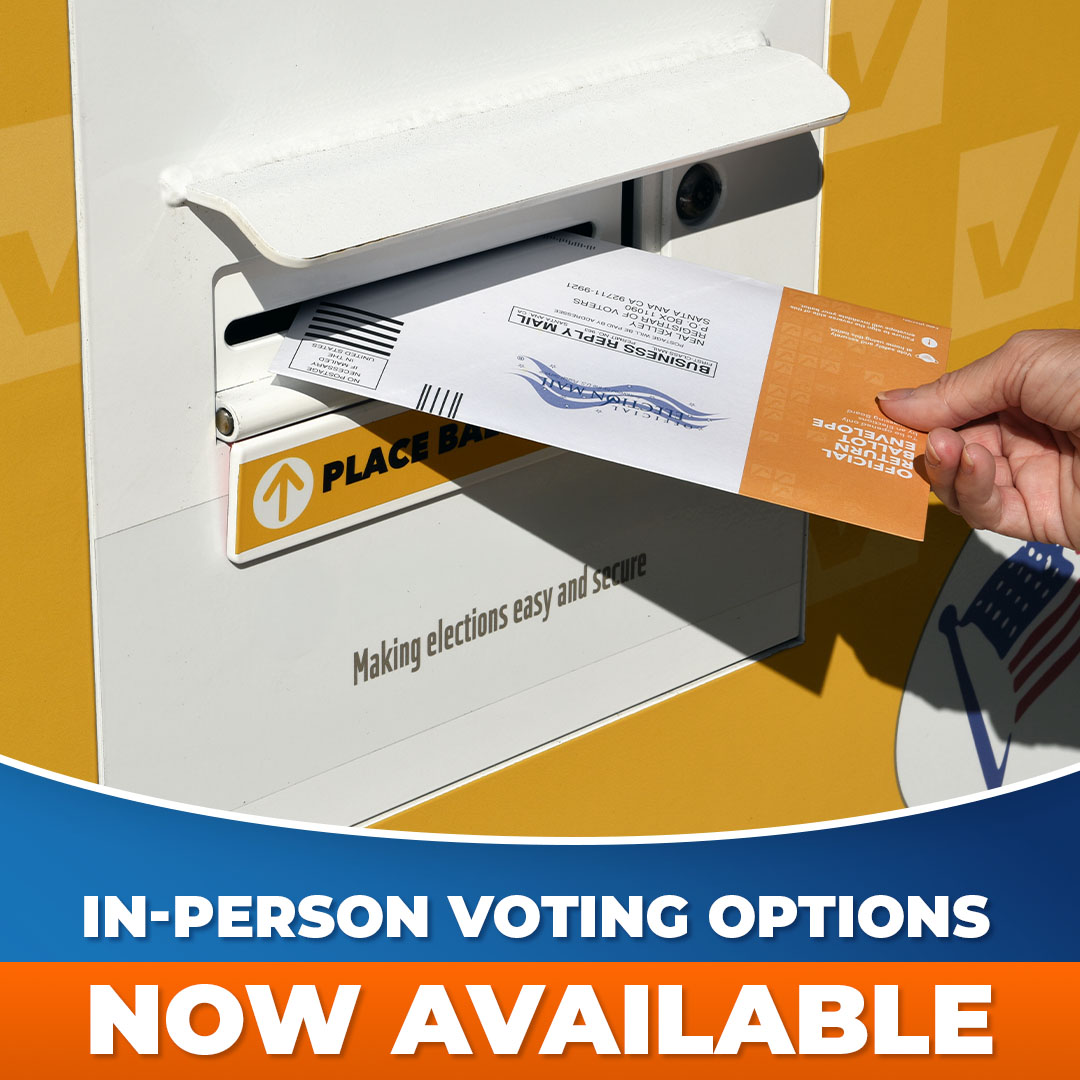 A hand inserts a ballot return envelope into an official ballot drop box beneath the text "In-Person Voting Options Now Available."